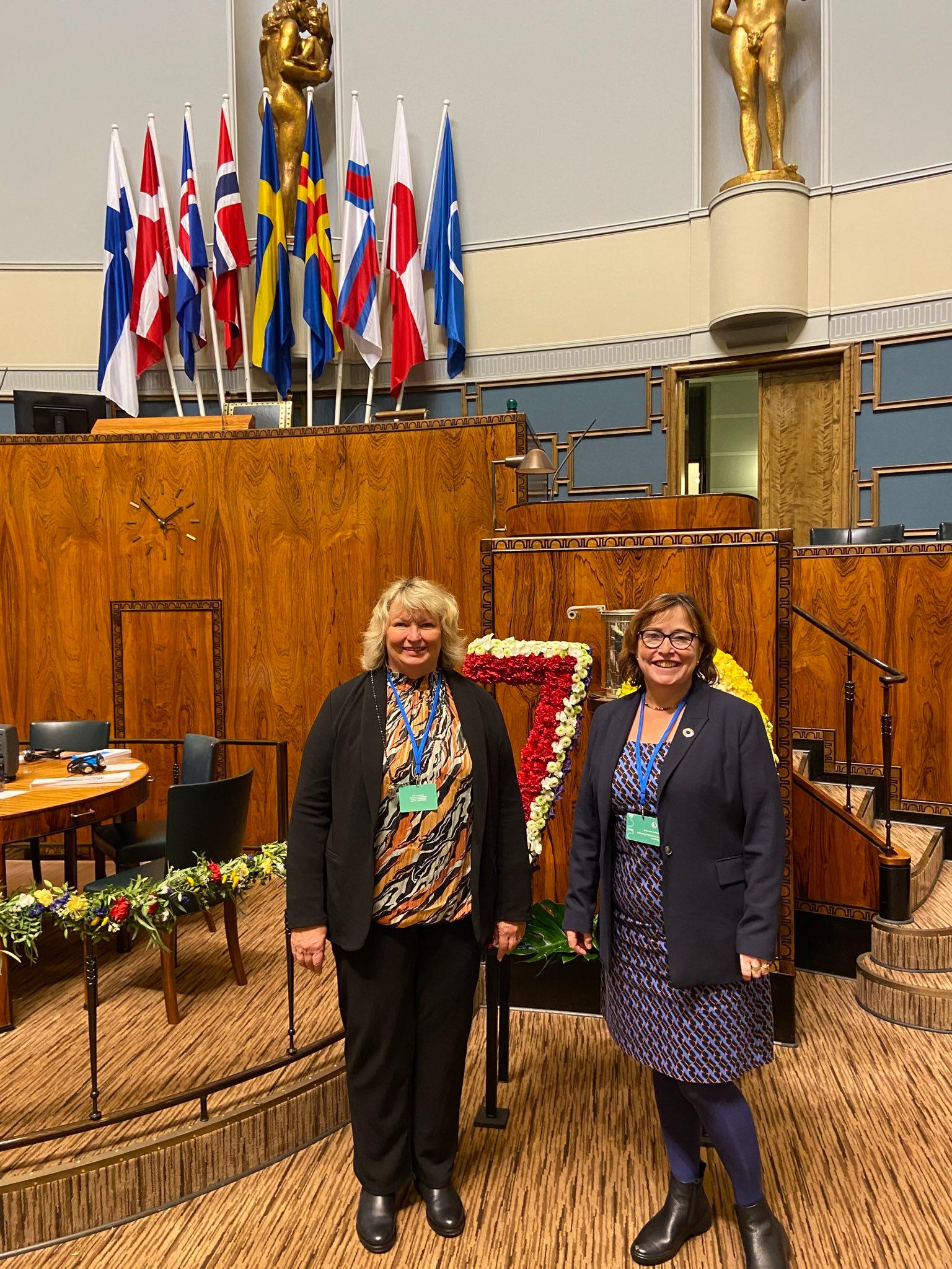 Foto von Eka von Kalben und Jette Waldinger-Thiering im Parlament in Helsinki. Im Hintergrund sieht man das Podium mit den skandinavischen Flaggen.