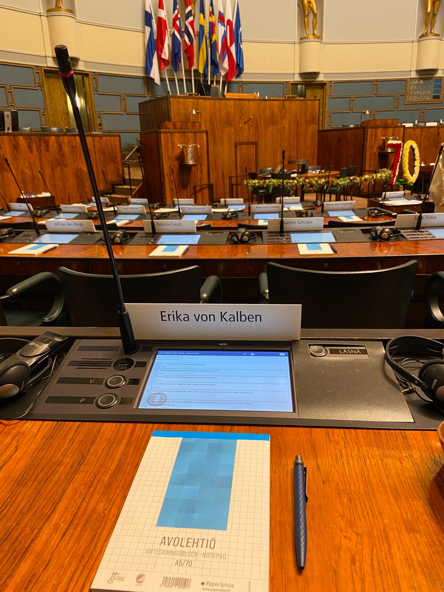 Foto von Eka von Kalbens Sitzplatz im leeren Parlament. Es gibt ein Namensschild, ein MIkro, ein eingebautes Tablet und einen Notizblock mit Kugelschreiber auf dem Tisch. Im Hintergrund sieht man das Präsidium mit den skandinavischen Flaggen.